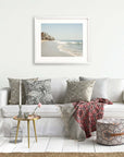 A cozy living room setup with a white sofa adorned with various patterned cushions, a throw blanket, a small round table with flowers, a cage-like decor item, and Offley Green's Malibu Beach House Print, 'Ocean View' wall art.