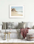 A cozy living room with a white couch adorned with decorative pillows, a framed Offley Green California Malibu Print, 'Point Dume' landscape on the wall, a small table with a vase next to the sofa, a blanket, and a pattern.