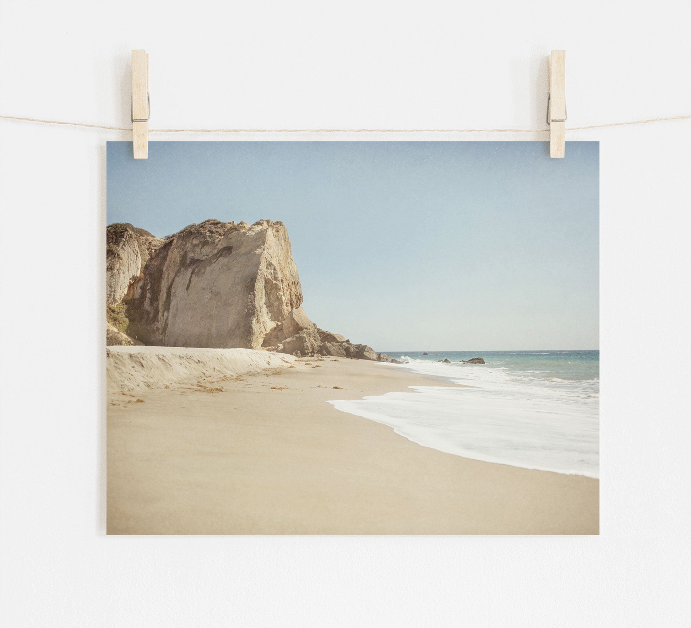 A photo of Offley Green's California Malibu Print, 'Point Dume' with waves gently lapping the shore and a large rocky outcrop to the left. The picture is pinned to a wall by wooden clothespins.