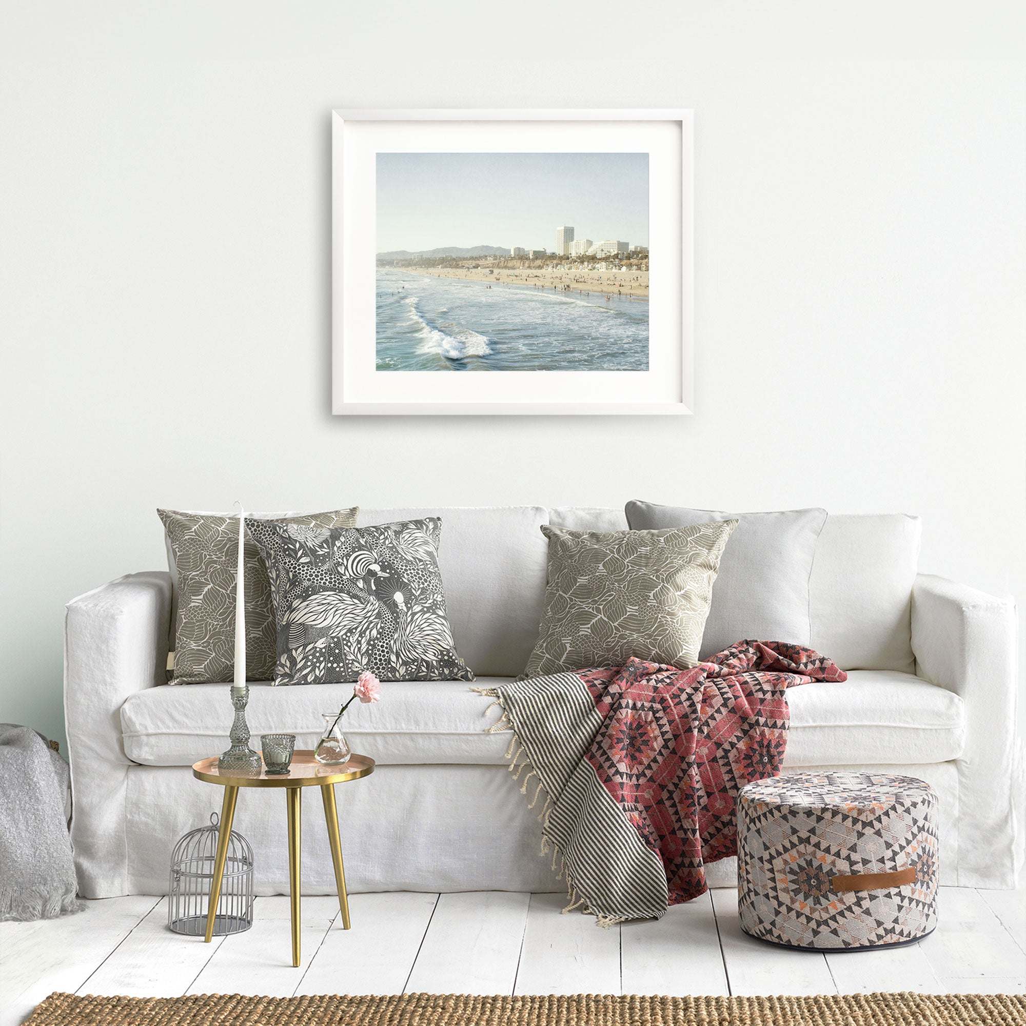 A cozy living room featuring a white sofa with decorative pillows, a throw blanket, and a small side table. Offley Green's 'Santa Monica Seaside' print hangs above the sofa.