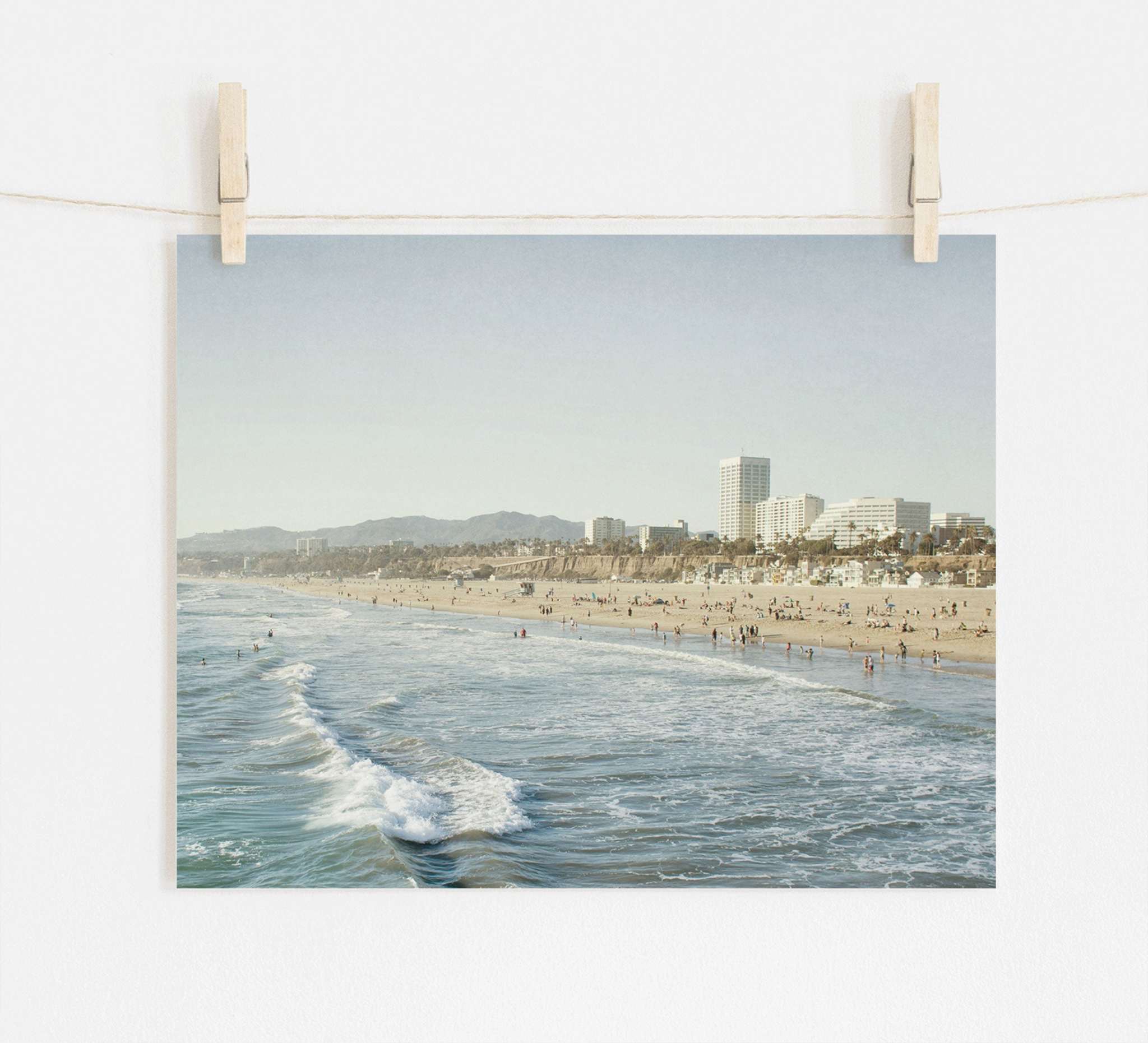 A photograph of a bustling beach scene with waves gently lapping the shore, pinned up by two wooden pegs against a plain background on archival photographic paper. The city skyline is visible in the distance. Offley Green's Santa Monica Print, 'Santa Monica Seaside'.