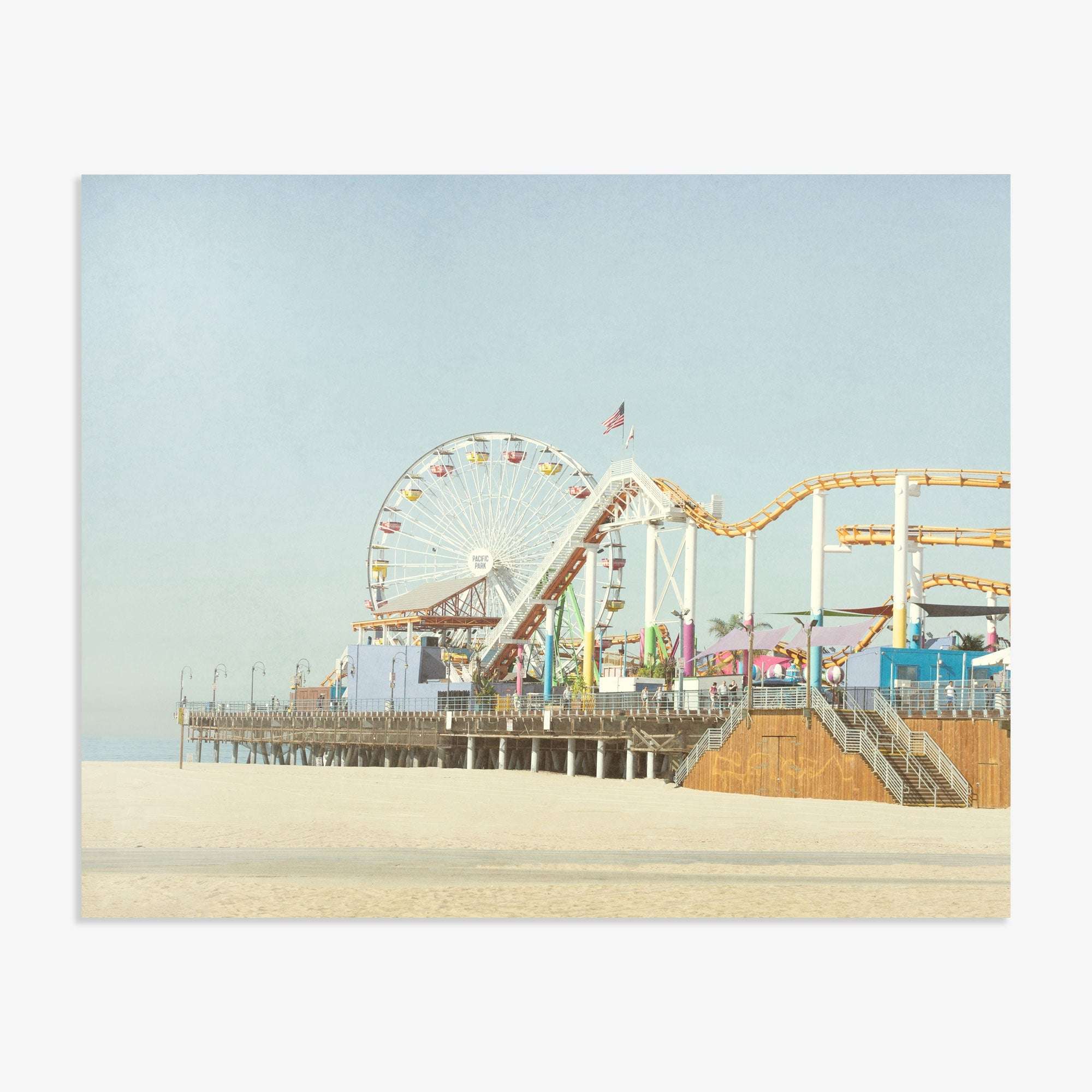 A seaside amusement park featuring a ferris wheel and roller coaster on the Santa Monica Pier, set against a clear sky. The sandy beach in the foreground is empty and expansive. Offley Green's California Print, 'Santa Monica Pier'.