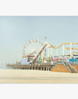 A seaside amusement park featuring a ferris wheel and roller coaster on the Santa Monica Pier, set against a clear sky. The sandy beach in the foreground is empty and expansive. Offley Green's California Print, 'Santa Monica Pier'.