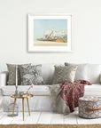 A cozy living room with a white sofa adorned with patterned pillows, a red throw blanket, a small round table, and a birdcage decor item, featuring a framed painting of the Offley Green California Print, 'Santa Monica Pier'