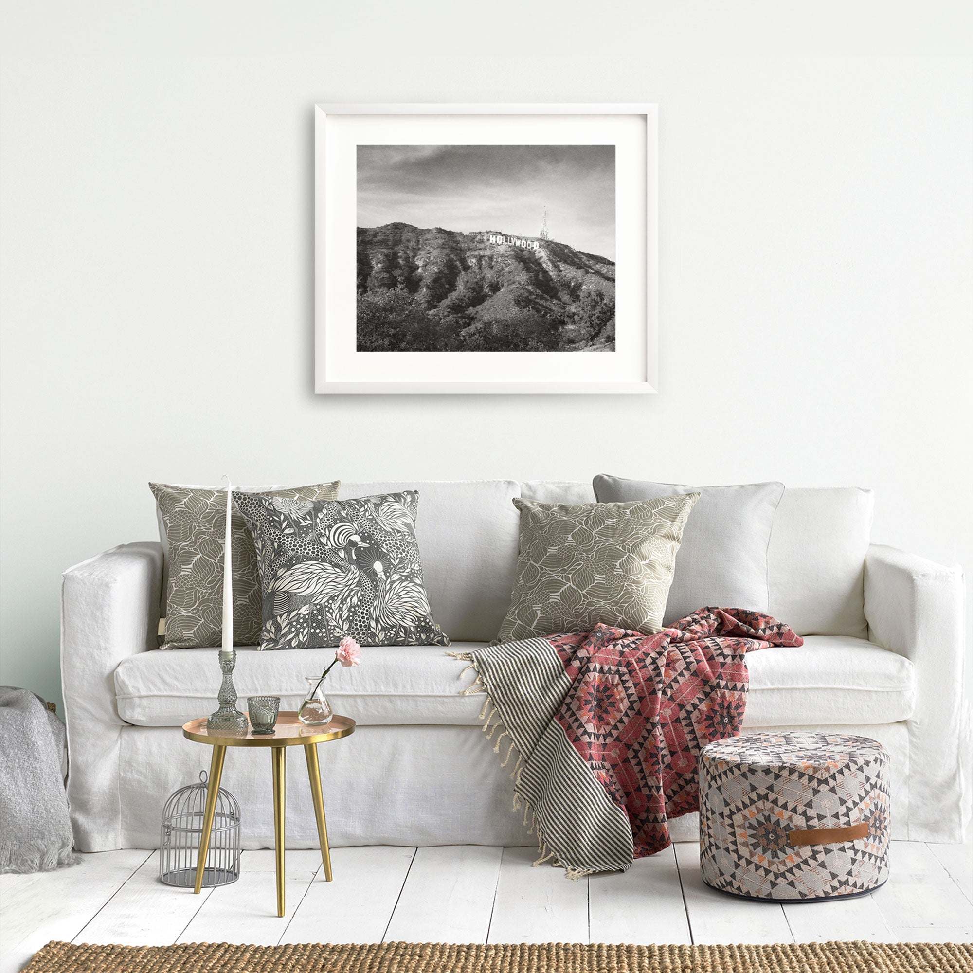 A cozy living room featuring a white sofa adorned with patterned pillows, a red and grey throw blanket, and a small round gold side table with books and a flower vase, against a wall with Offley Green's Hollywood Sign Black and White Vintage Print, 'Old Hollywood'.