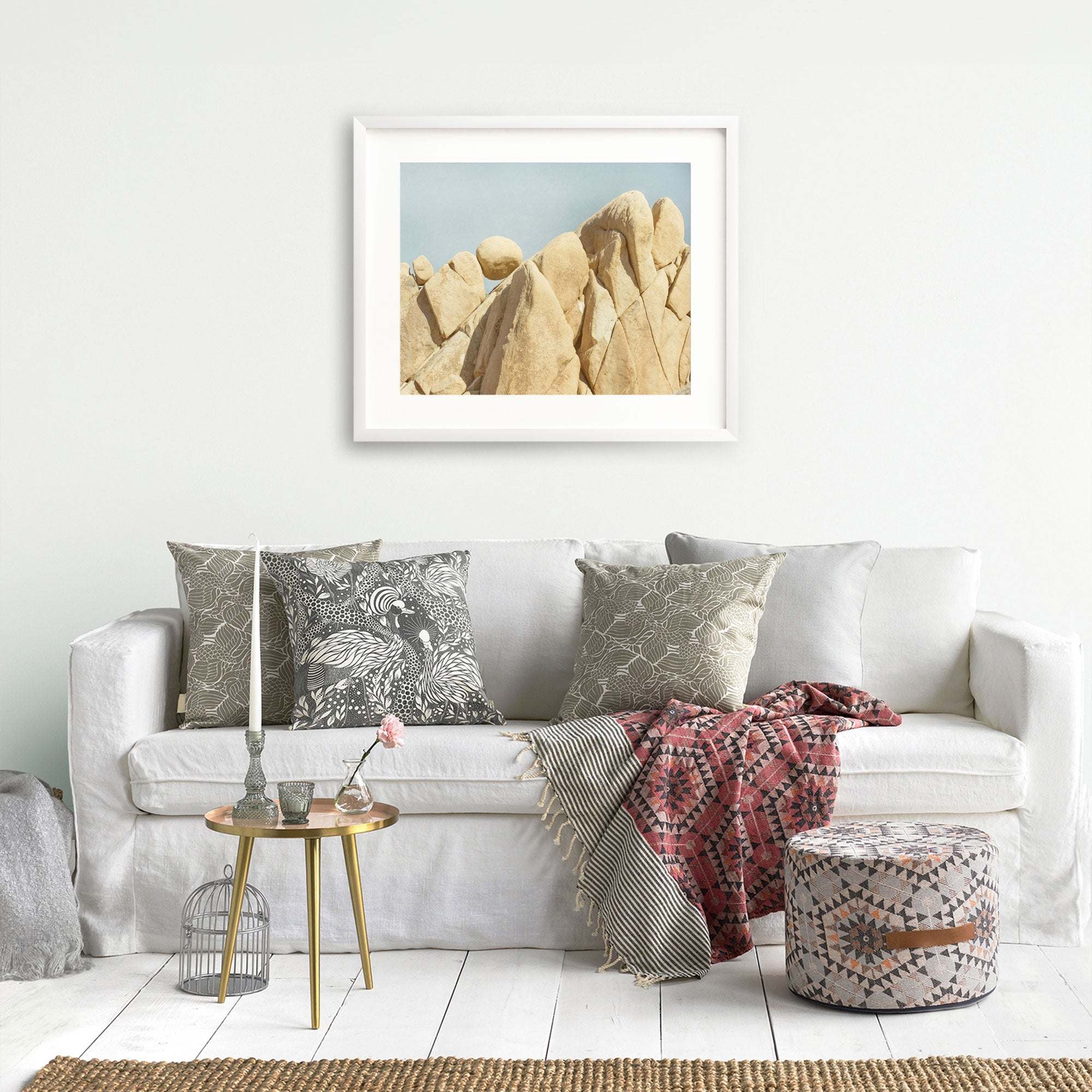 A cozy living room with a white sofa adorned with patterned cushions, a red patterned throw, a small coffee table with books and a vase, an unframed Offley Green print of Joshua Tree National Park 'Rock Formations'.