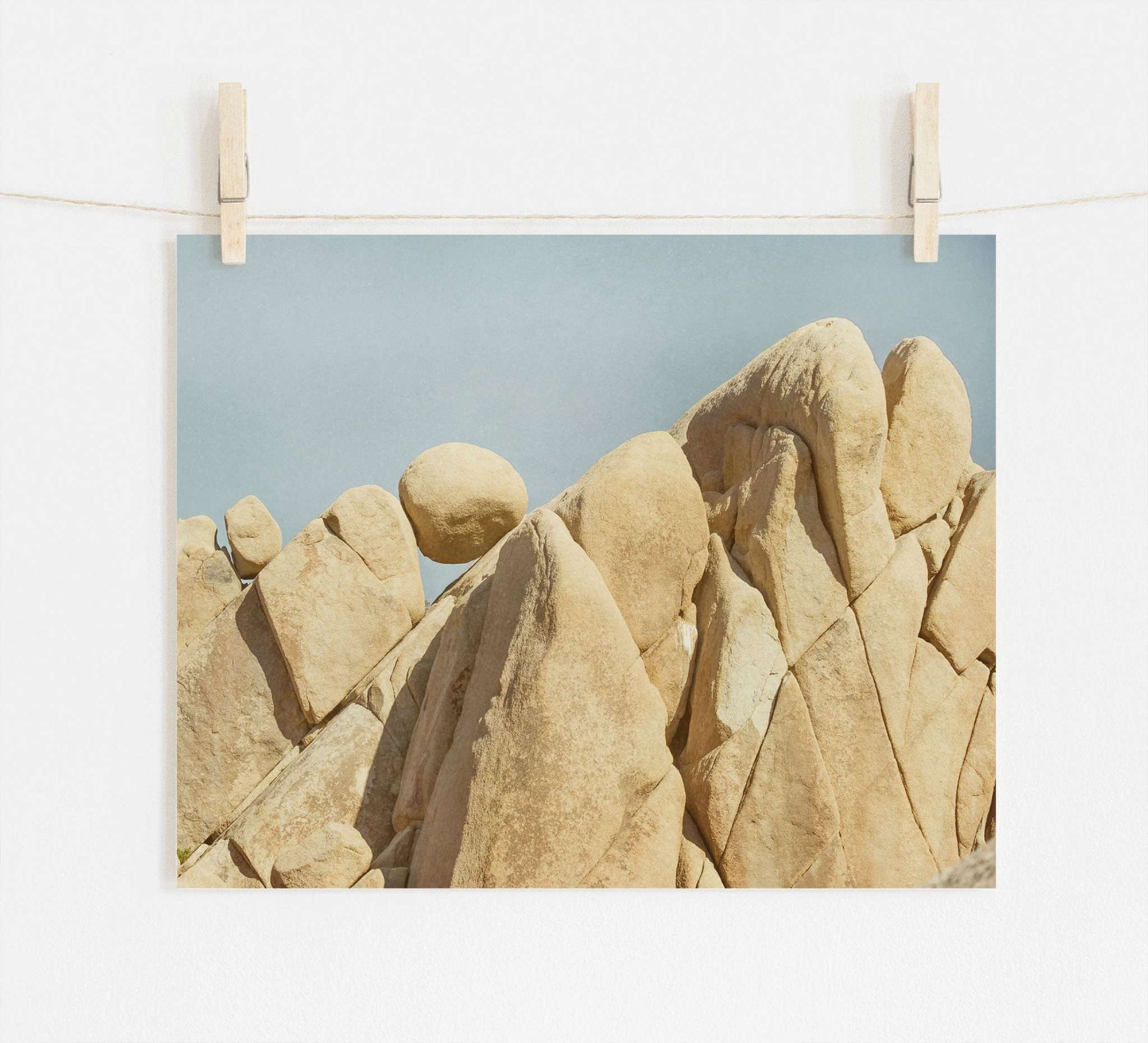 A photograph of rugged, beige rocks in Joshua Tree National Park against a clear blue sky, clipped to a string with two wooden clothespins, displayed against a white wall. Offley Green's Joshua Tree Print, 'Rock Formations.'