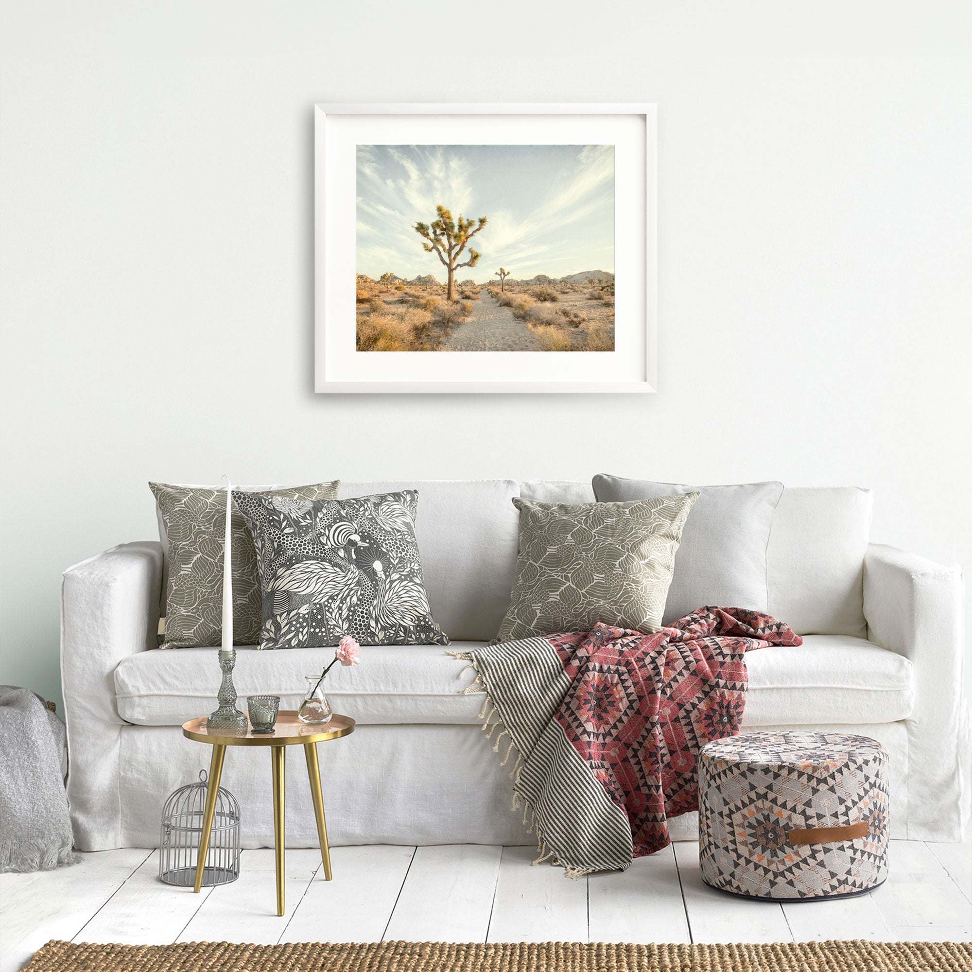 A well-decorated living room with a white couch adorned with patterned pillows, a stylish throw, and a small table. A framed Offley Green 'Path to Joshua' print hangs above the couch.