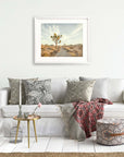 A well-decorated living room with a white couch adorned with patterned pillows, a stylish throw, and a small table. A framed Offley Green 'Path to Joshua' print hangs above the couch.