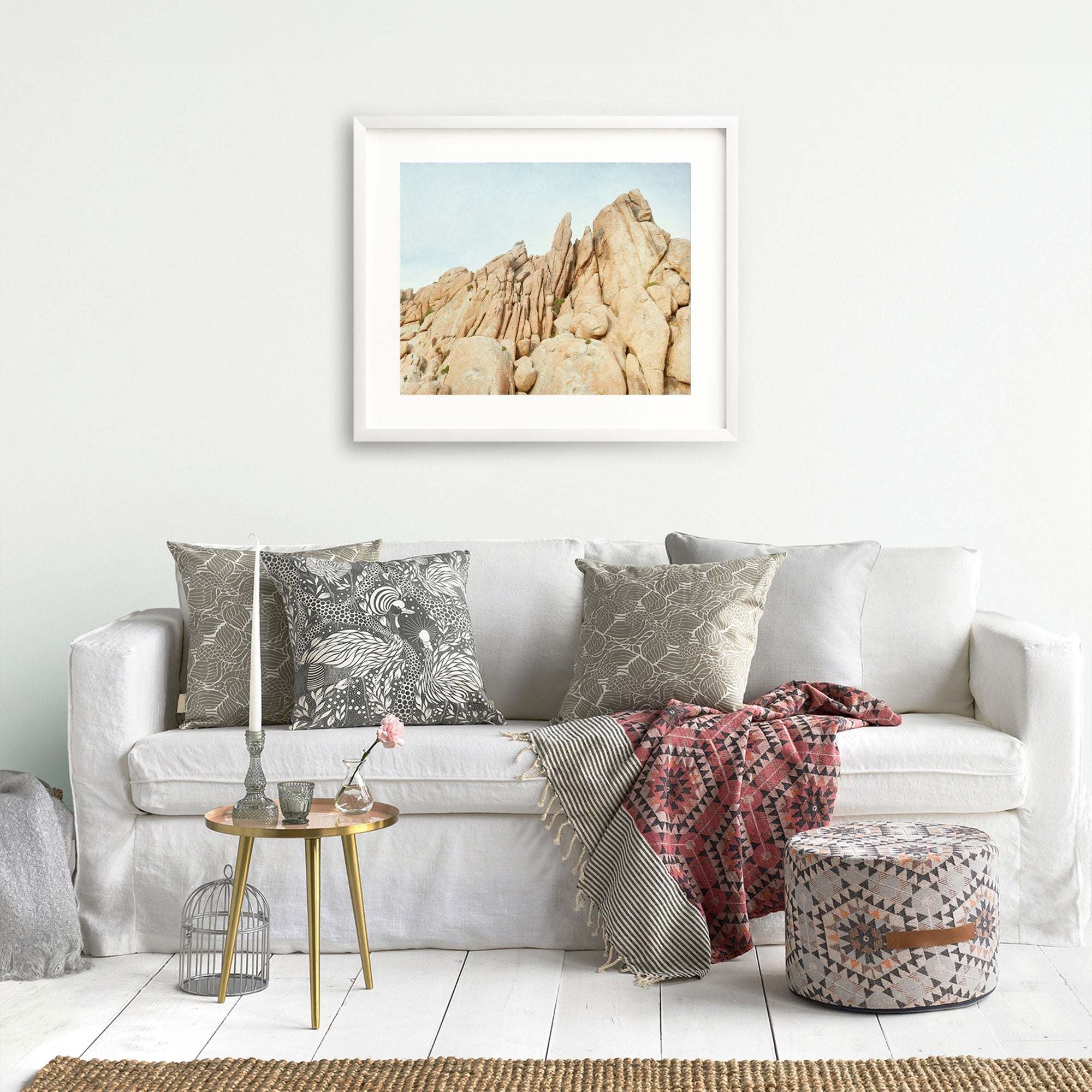 A cozy living room featuring a white sofa adorned with decorative gray pillows, a red patterned throw blanket, a small round table with a plant, a birdcage, and unframed prints of Offley Green's Joshua Tree Print, 'Joshua Rocks'.