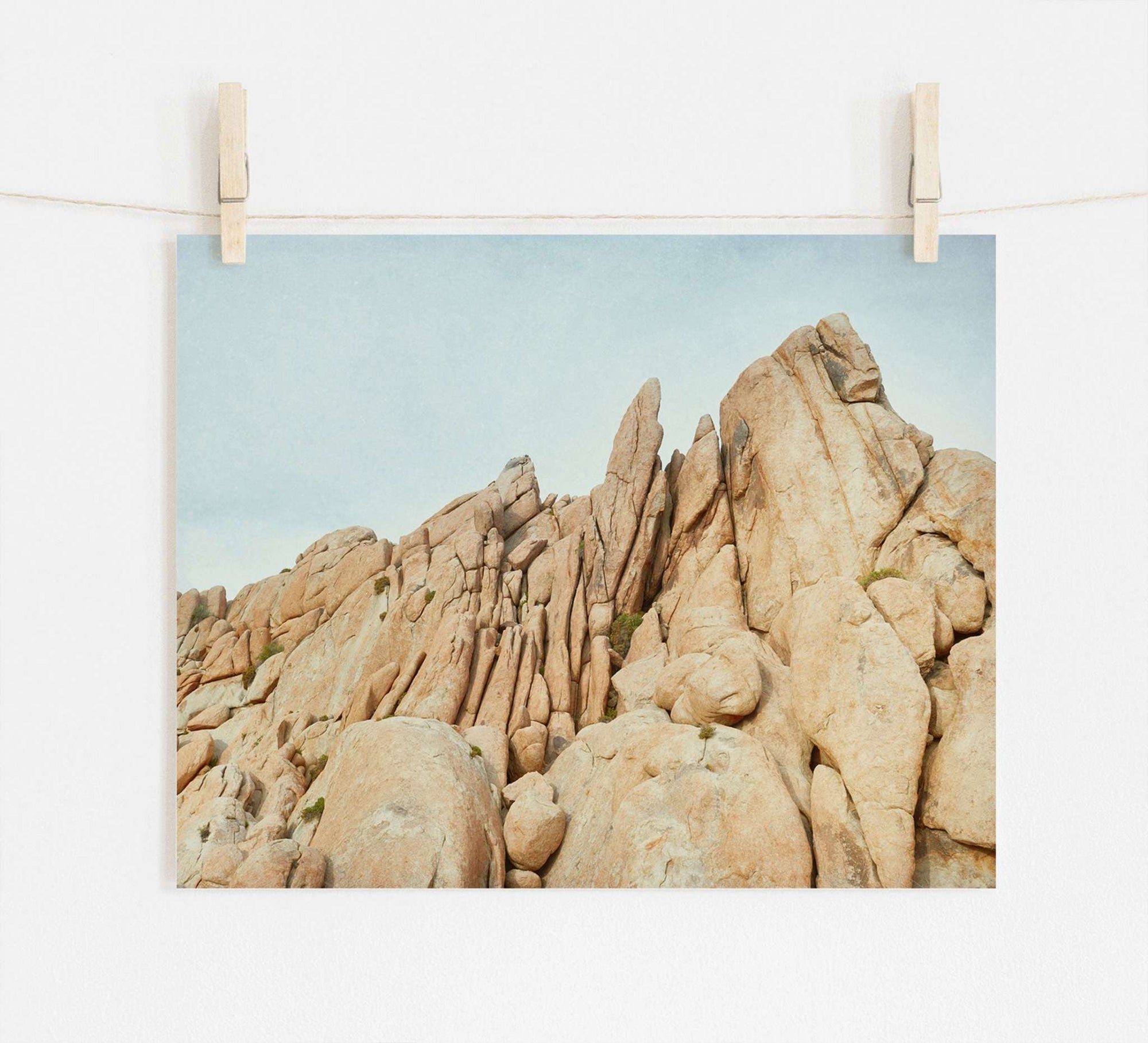 A photo of rugged, rocky cliffs with pointed formations, displayed hanging from two wooden clothespins on a string against a white wall, printed on archival photographic paper. 
Replace with: Offley Green's Joshua Tree Print, 'Joshua Rocks'