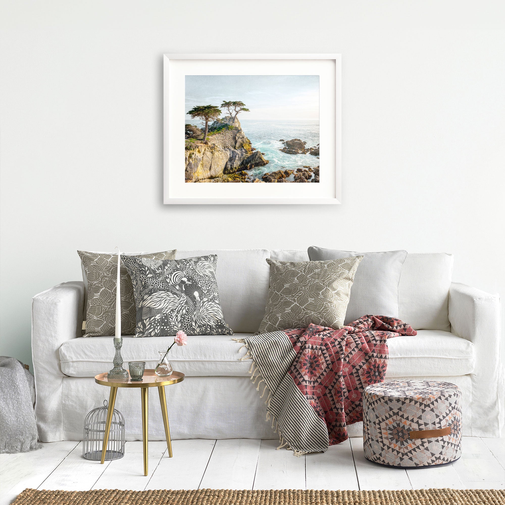 A cozy living room setup featuring a white sofa with decorative pillows, a red throw, a small round table with unframed prints and books, and an Offley Green California Coastal Print, 'Lone Cypress' painting on the wall.