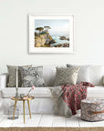 A cozy living room setup featuring a white sofa with decorative pillows, a red throw, a small round table with unframed prints and books, and an Offley Green California Coastal Print, 'Lone Cypress' painting on the wall.