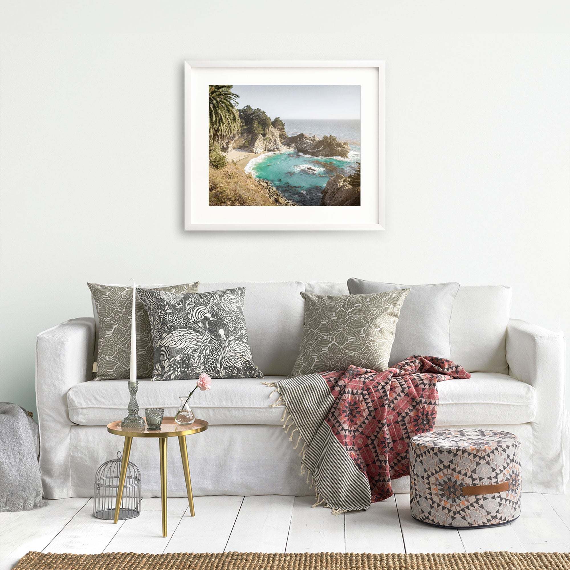 A cozy living room with a white sofa adorned with assorted patterned throw pillows and a colorful blanket. A small round table with decorative items sits beside the sofa. On the wall behind the sofa hangs a framed Offley Green Big Sur Coastal Print, 'Julia Pffeifer,' reminiscent of Julia Pfeiffer Burns State Park, Big Sur.