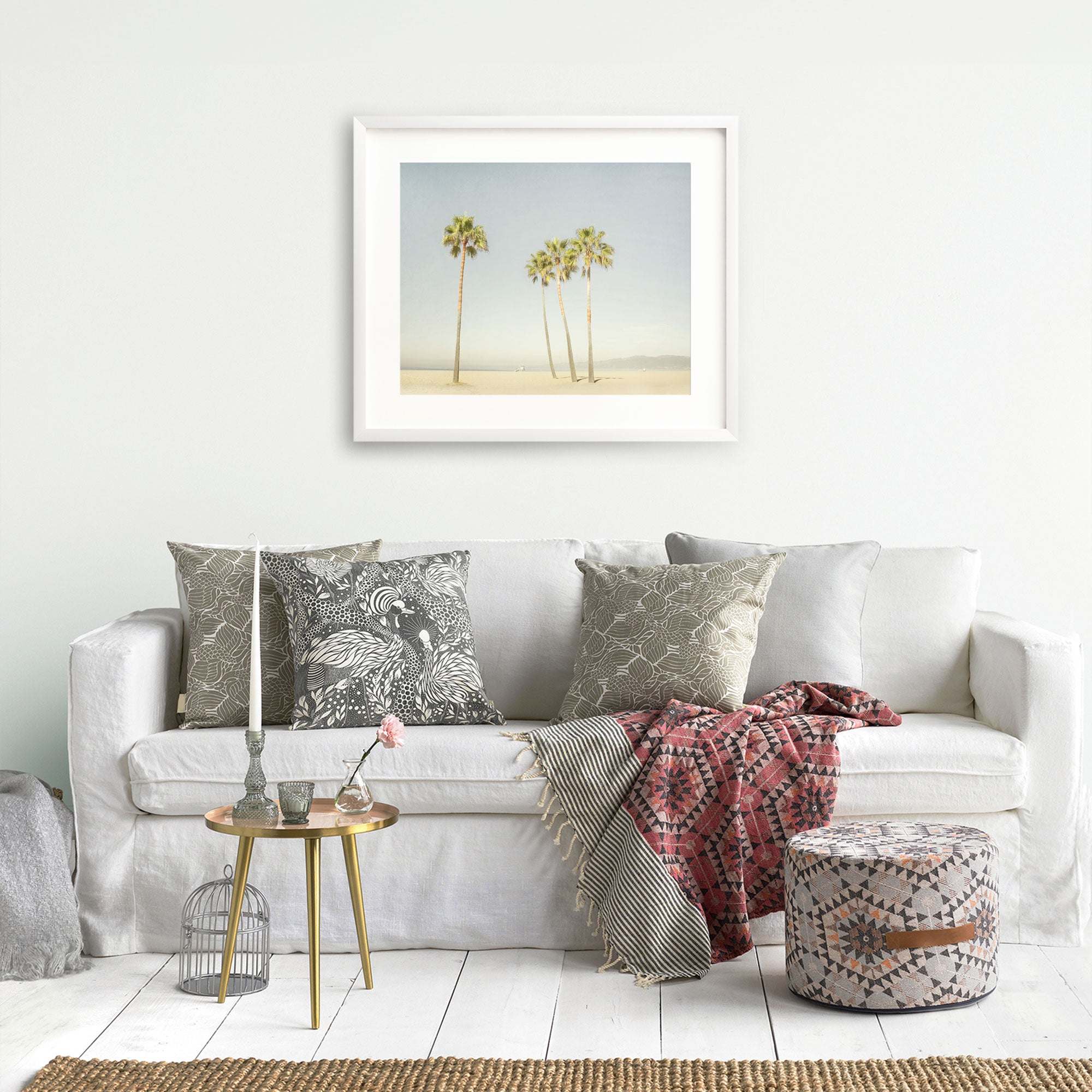 A cozy living room scene with a white sofa adorned with patterned cushions, a framed California Beach Palm Tree Print from Offley Green on the wall, a small gold table with a plant and books, a patterned ottoman, and a decorative birdcage.