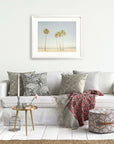 A cozy living room scene with a white sofa adorned with patterned cushions, a framed California Beach Palm Tree Print from Offley Green on the wall, a small gold table with a plant and books, a patterned ottoman, and a decorative birdcage.