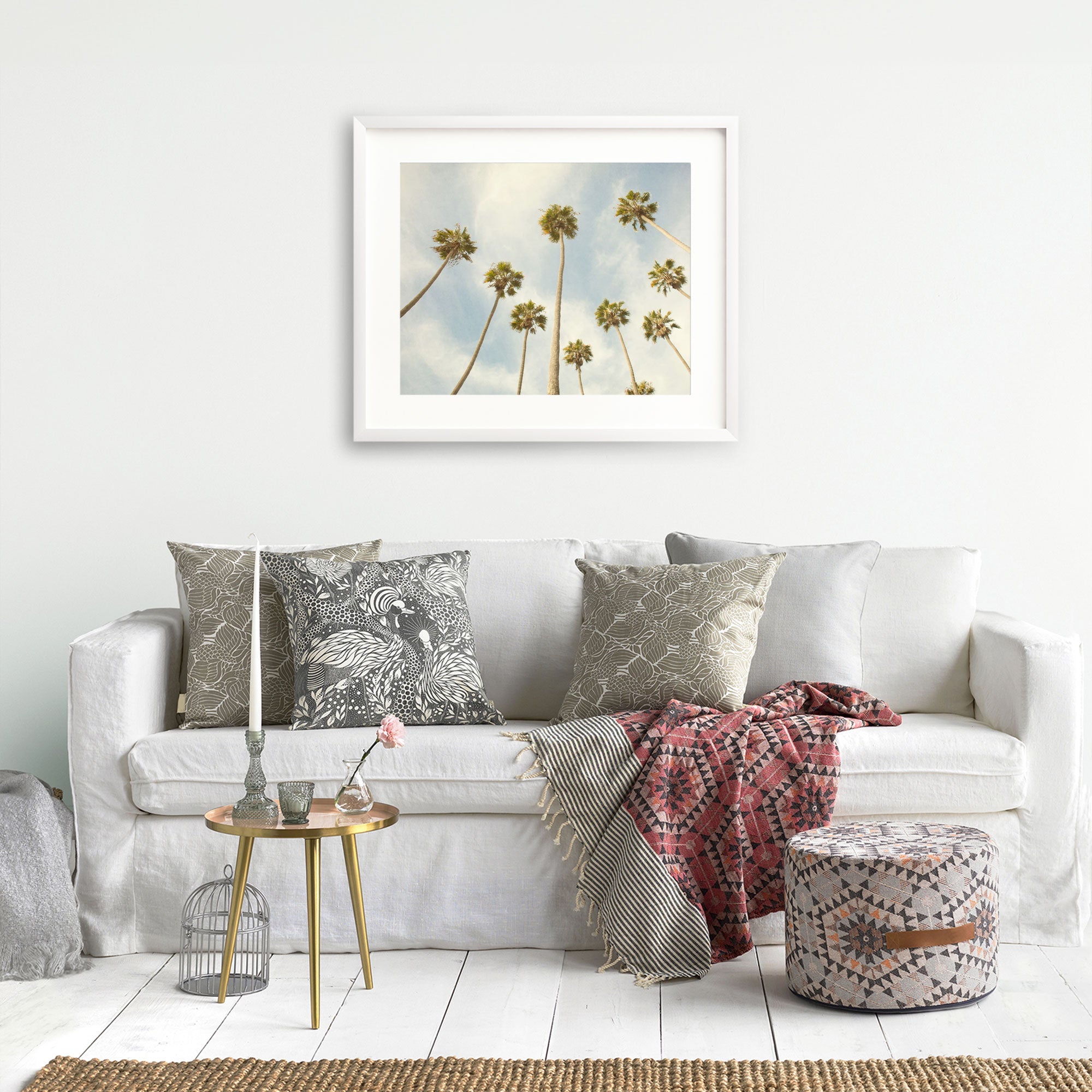 A cozy living room with a white sofa adorned with patterned pillows and a red throw blanket, a small round table, a decorative cage, and an Offley Green 'Reach for the Palms' palm tree print depicting a California beach scene hanging on the wall.