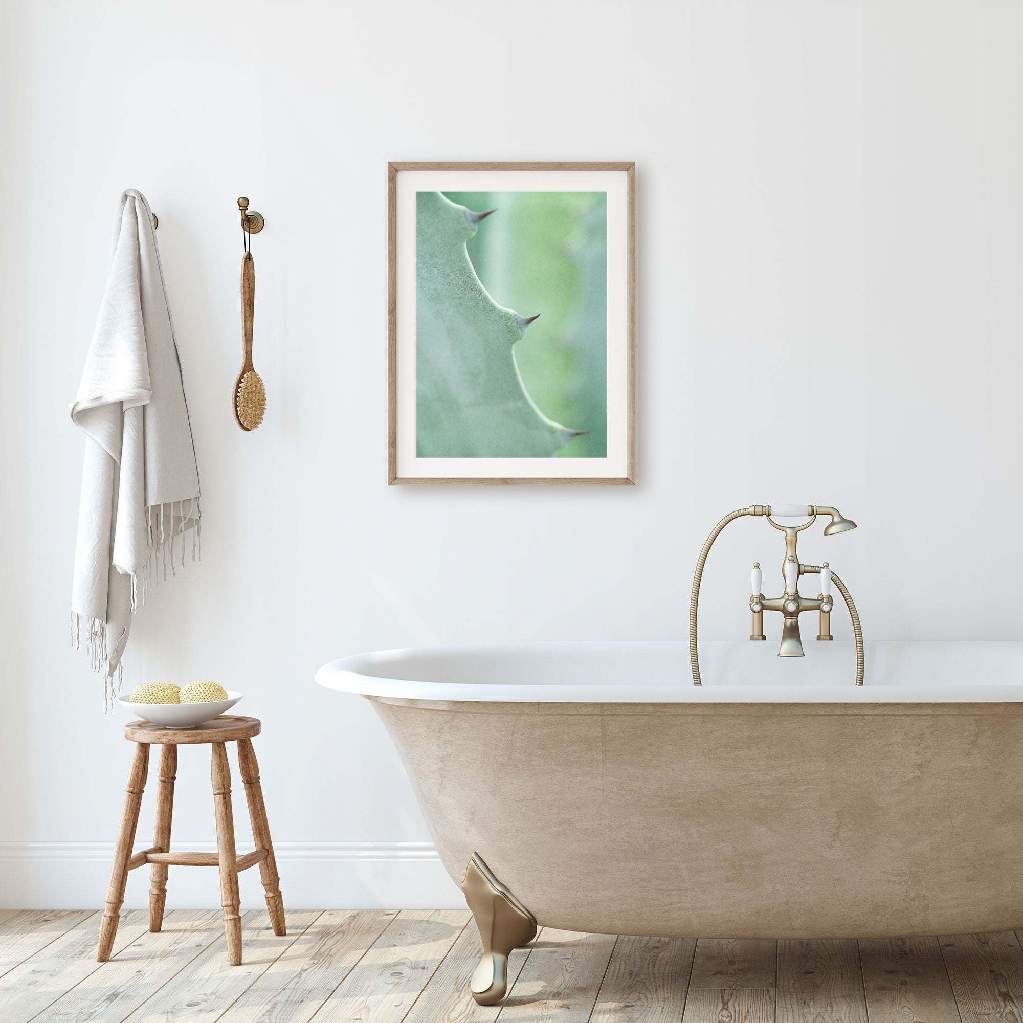 A minimalistic bathroom featuring a freestanding bathtub with golden claw feet, a wooden stool with bath items, a towel hanging on the wall, and Offley Green's 'Aloe Vera Spikes II' unframed print above the tub.