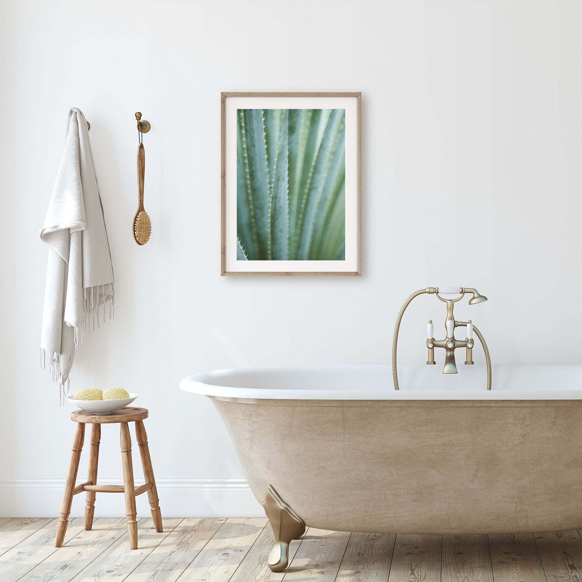 A minimalist bathroom with a freestanding bathtub, a stool holding bath accessories, and an unframed &#39;Strands and Spikes II&#39; desert plants photograph by Offley Green on the wall. A towel hangs beside a wooden brush.