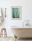A minimalist bathroom with a freestanding bathtub, a stool holding bath accessories, and an unframed 'Strands and Spikes II' desert plants photograph by Offley Green on the wall. A towel hangs beside a wooden brush.