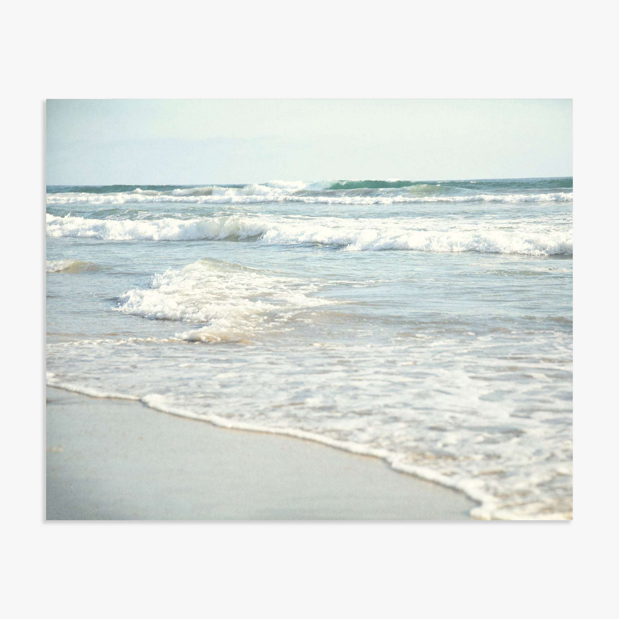 Ocean waves roll onto Carlsbad State Beach, capturing the dynamic movement of water with a backdrop of a hazy, overcast sky featuring the Coastal Beach Print in California 'Surf and Sun' by Offley Green.