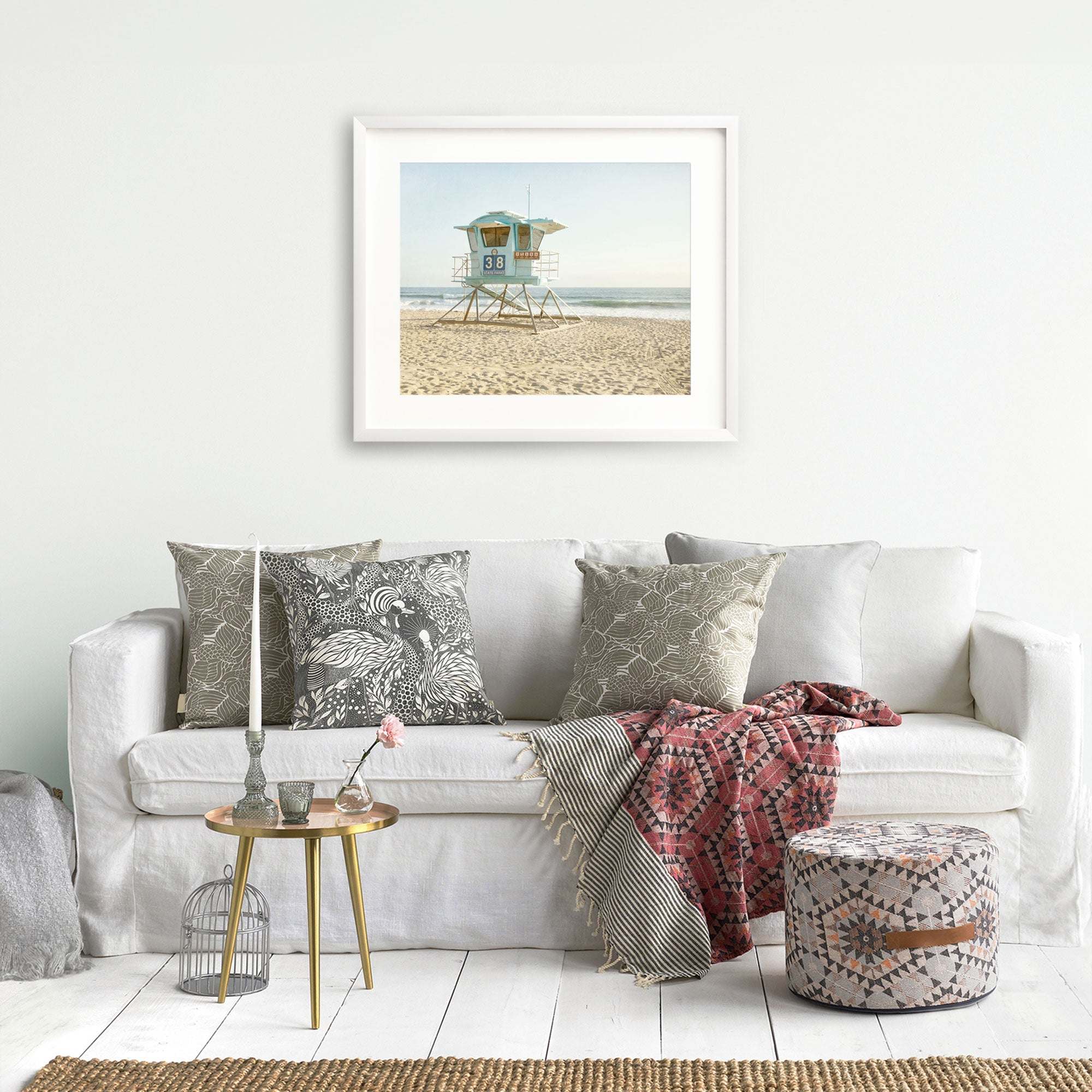 A cozy living room featuring a white sofa adorned with patterned cushions, a small table with decor, a wicker ottoman, and a framed print of the California Coastal Print, 'Carlsbad Lifeguard Tower' by Offley Green hanging on the wall.
