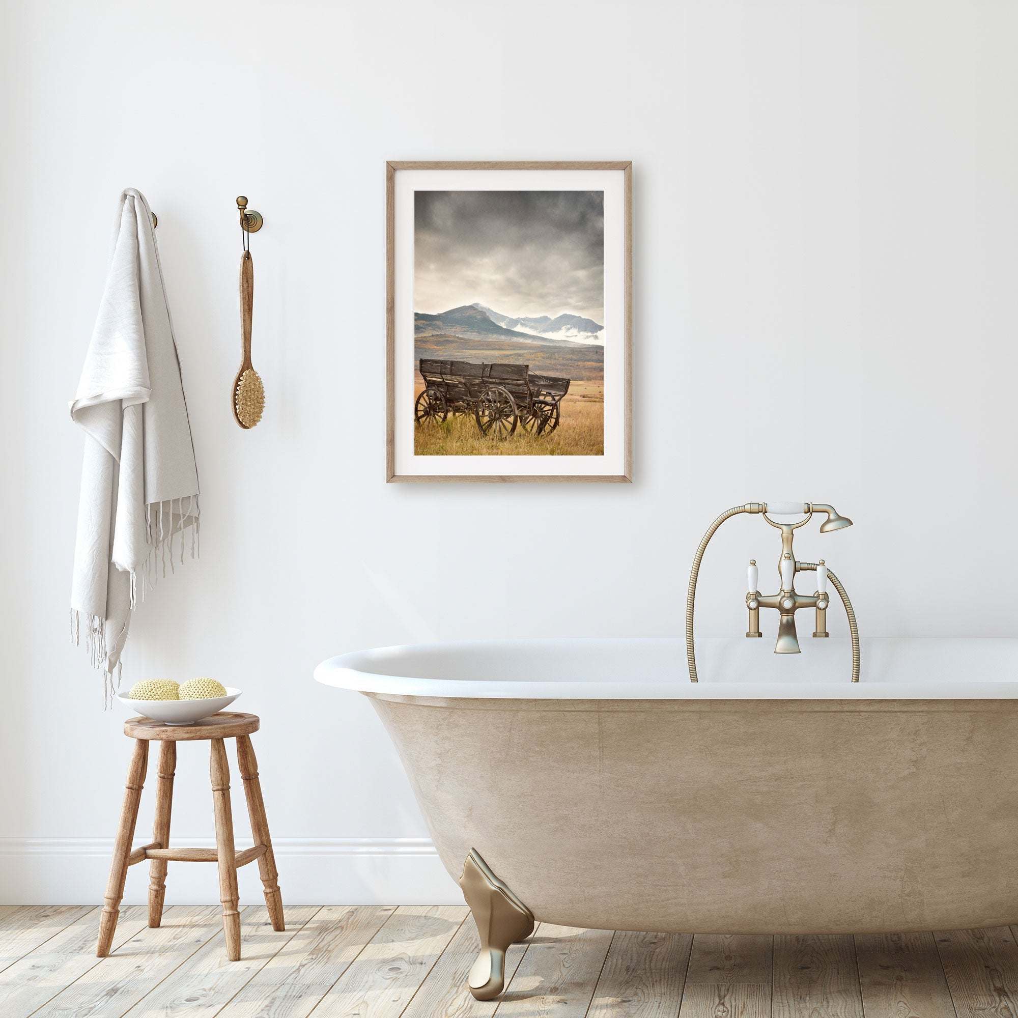 A bathroom exudes rustic charm with a vintage-style clawfoot bathtub and faucet, a wooden stool with towels and soap, a towel on a hook, and Offley Green's Rustic Farmhouse Wall Art, 'Old Wagon', adorning the wall.
