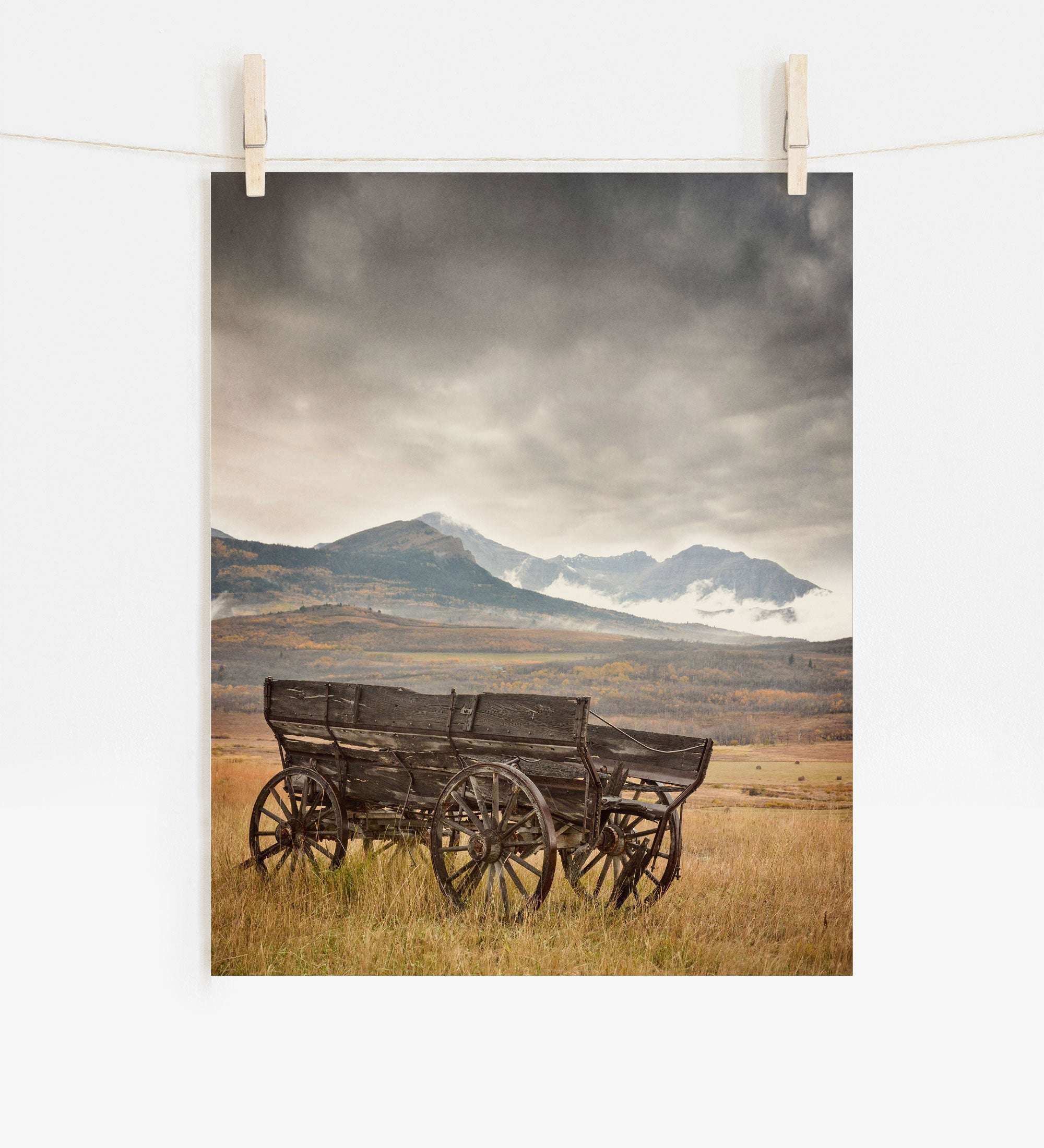 The "Rustic Farmhouse Wall Art, 'Old Wagon'" by Offley Green depicts a weathered wooden wagon in an open field under a cloudy sky, with misty mountains in the distance. This serene image is expertly printed on photographic paper and clipped to a white wall with clothespins.