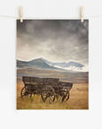 The "Rustic Farmhouse Wall Art, 'Old Wagon'" by Offley Green depicts a weathered wooden wagon in an open field under a cloudy sky, with misty mountains in the distance. This serene image is expertly printed on photographic paper and clipped to a white wall with clothespins.