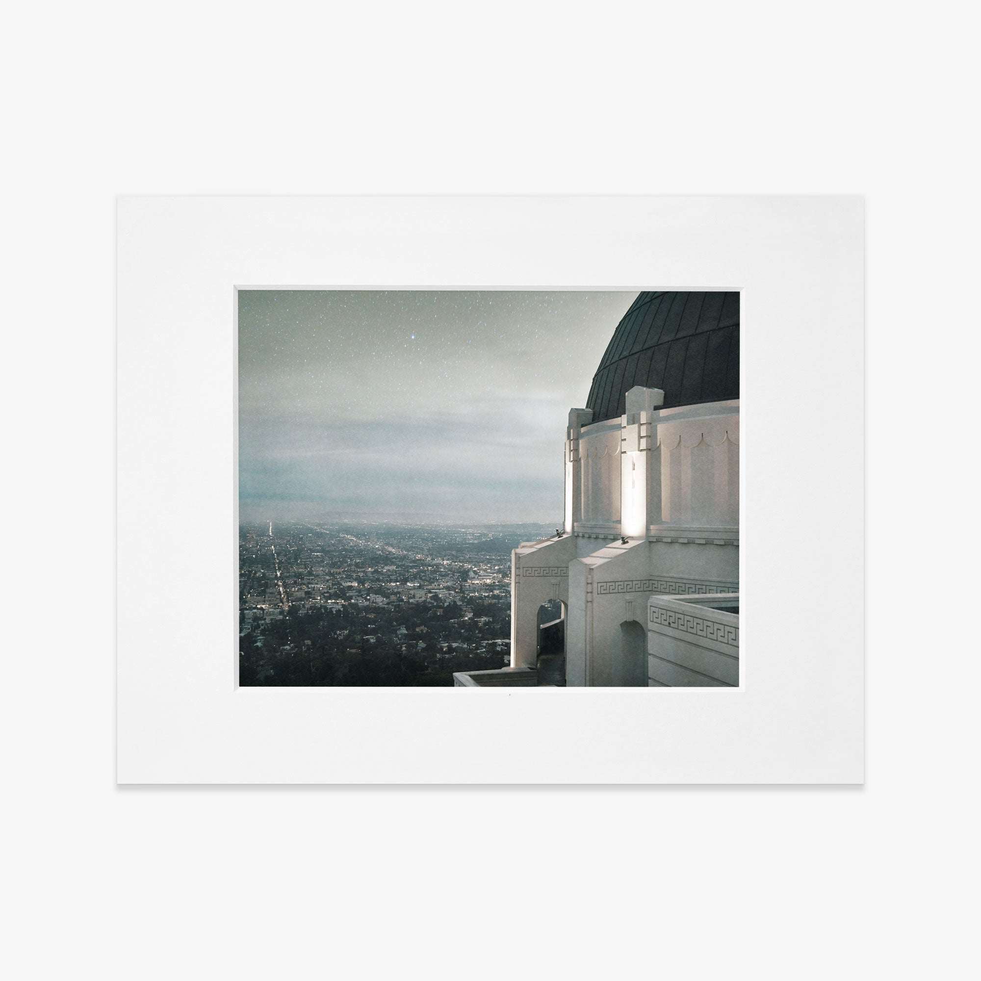 A nighttime photograph framed in white, showcasing the Los Angeles city view from Offley Green&#39;s Griffith Observatory Print, &#39;The Sky At Night&#39;, with twinkling lights under a starry sky.
