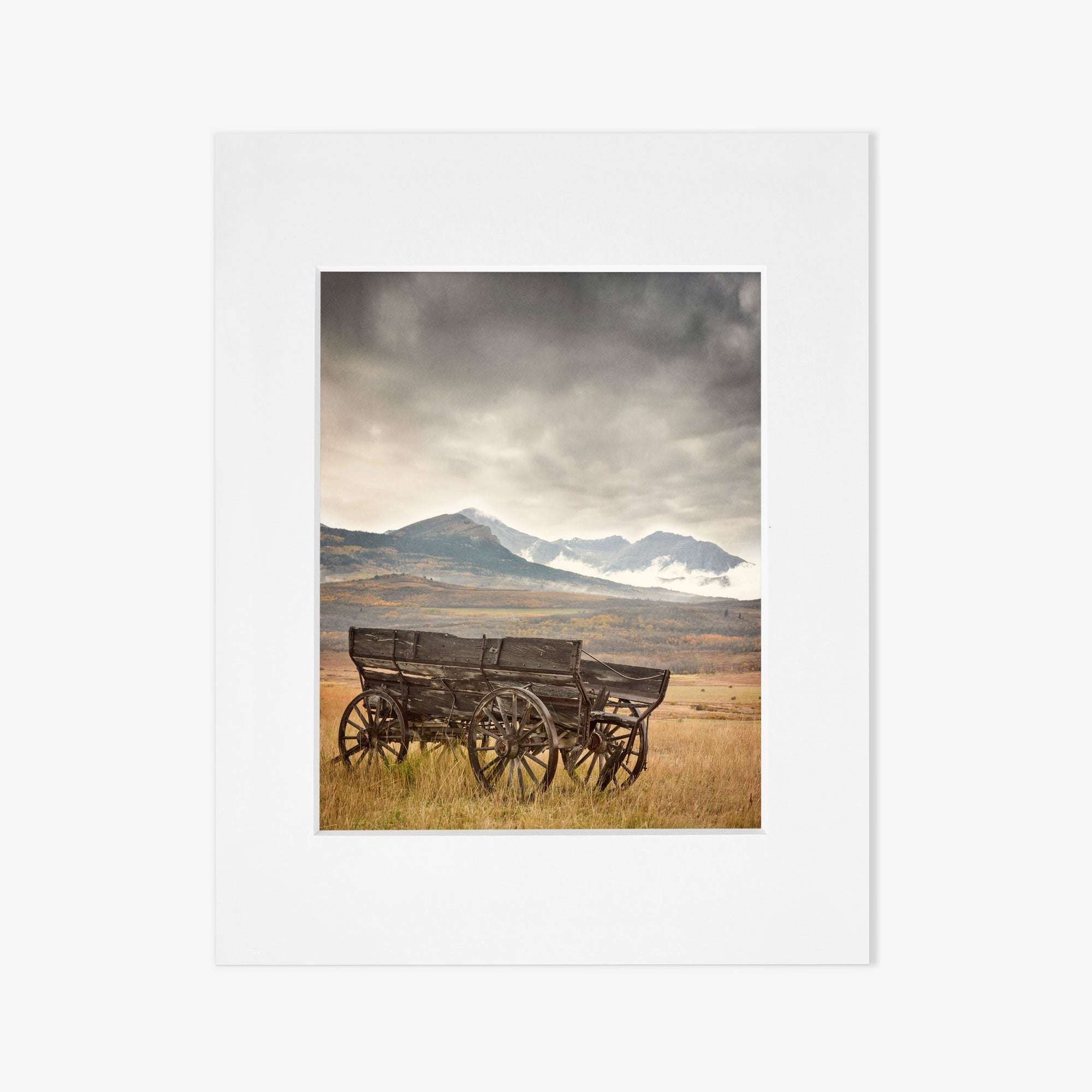 Offley Green's 'Old Wagon' Rustic Farmhouse Wall Art features a scenic view of an antique wooden wagon with large wheels on a grassy field, set against snow-capped mountains under a cloudy sky, capturing a nostalgic and tranquil landscape.