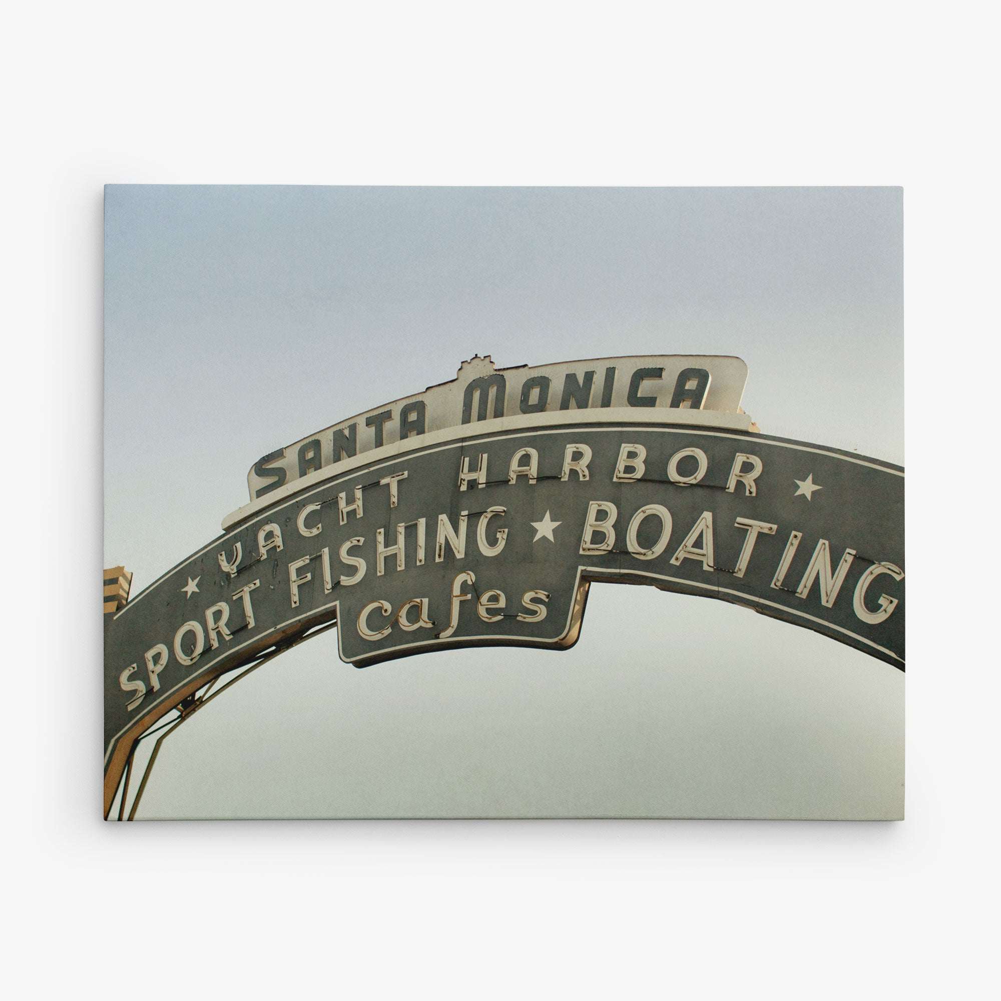 A vintage-style sign reading "Santa Monica Yacht Harbor Sport Fishing Boating Cafes" is set against a clear sky at the Santa Monica Pier. This classic piece of wall art, the Los Angeles California Canvas Wall Art, 'Santa Monica Pier Blues' by Offley Green, features arched text with stars separating the activities listed, evoking the charm of a summer Californian beach town.