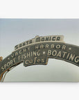 A vintage-style sign reading "Santa Monica Yacht Harbor Sport Fishing Boating Cafes" is set against a clear sky at the Santa Monica Pier. This classic piece of wall art, the Los Angeles California Canvas Wall Art, 'Santa Monica Pier Blues' by Offley Green, features arched text with stars separating the activities listed, evoking the charm of a summer Californian beach town.