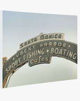 A vintage-style, sepia-toned image of an arched sign that reads "Santa Monica Yacht Harbor, Sport Fishing, Boating, Cafes" against a pale sky. The sign has neon lights and is iconic for marking the entrance to the Santa Monica Pier, capturing the essence of a summer Californian beach town as perfect wall art like Los Angeles California Canvas Wall Art, 'Santa Monica Pier Blues' by Offley Green.