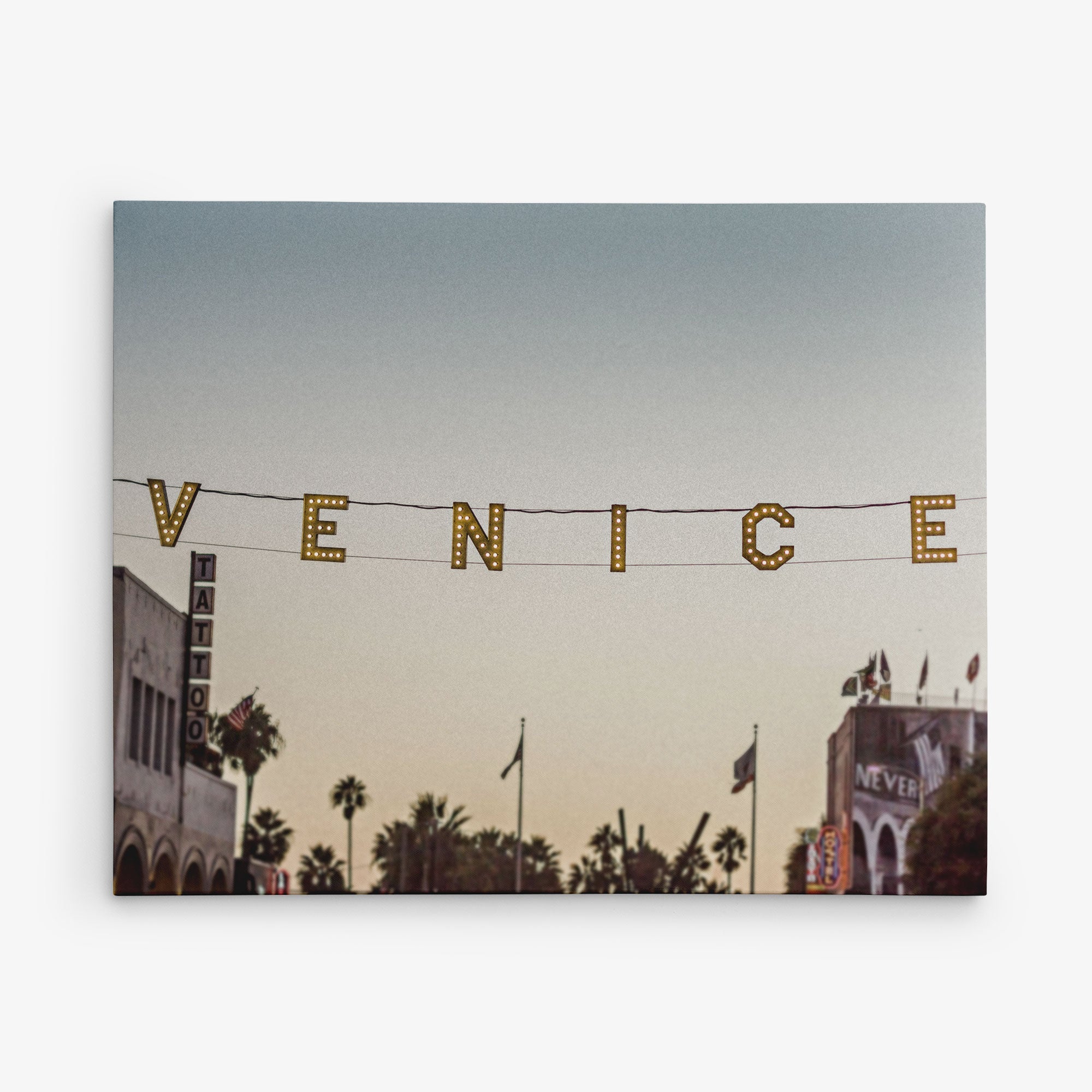A photo of the Venice sign in Venice Beach, California. The illuminated letters hang across the street at sunset, capturing the essence of this iconic locale. Palm trees and buildings, including a tattoo shop and a "NEVER" sign, add to the charm—perfect for Offley Green's 16x20 California Canvas Print (Choose from 10+ Designs).