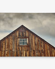 A rustic wooden barn with a weathered exterior and a small, single-pane window near the roof peak. The sky in the background is cloudy and gray, adding a moody atmosphere to the scene. This photograph, ready to hang as 30x40 Rustic Canvas Print (Choose from 10+ Designs), focuses on the top half of the barn by Offley Green.