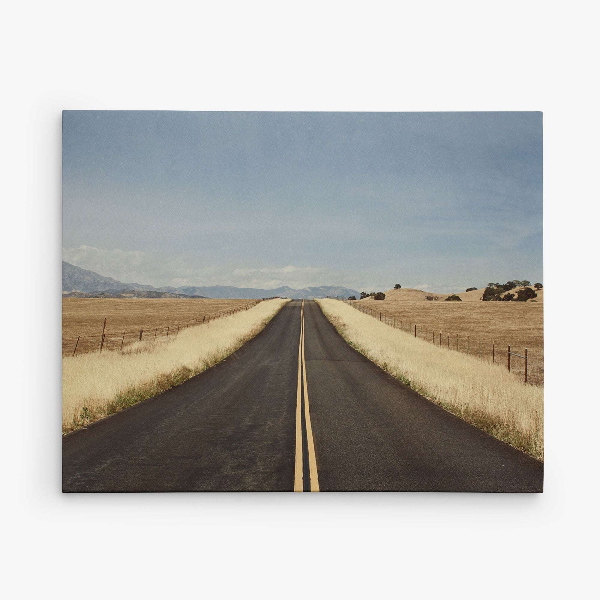 A straight, empty road with a double yellow line through a rural landscape evokes the essence of an Offley Green 8x10 Rustic Canvas Print. Dry grass flanks the road while distant hills stretch beneath a clear blue sky, reminiscent of timeless rustic print designs.