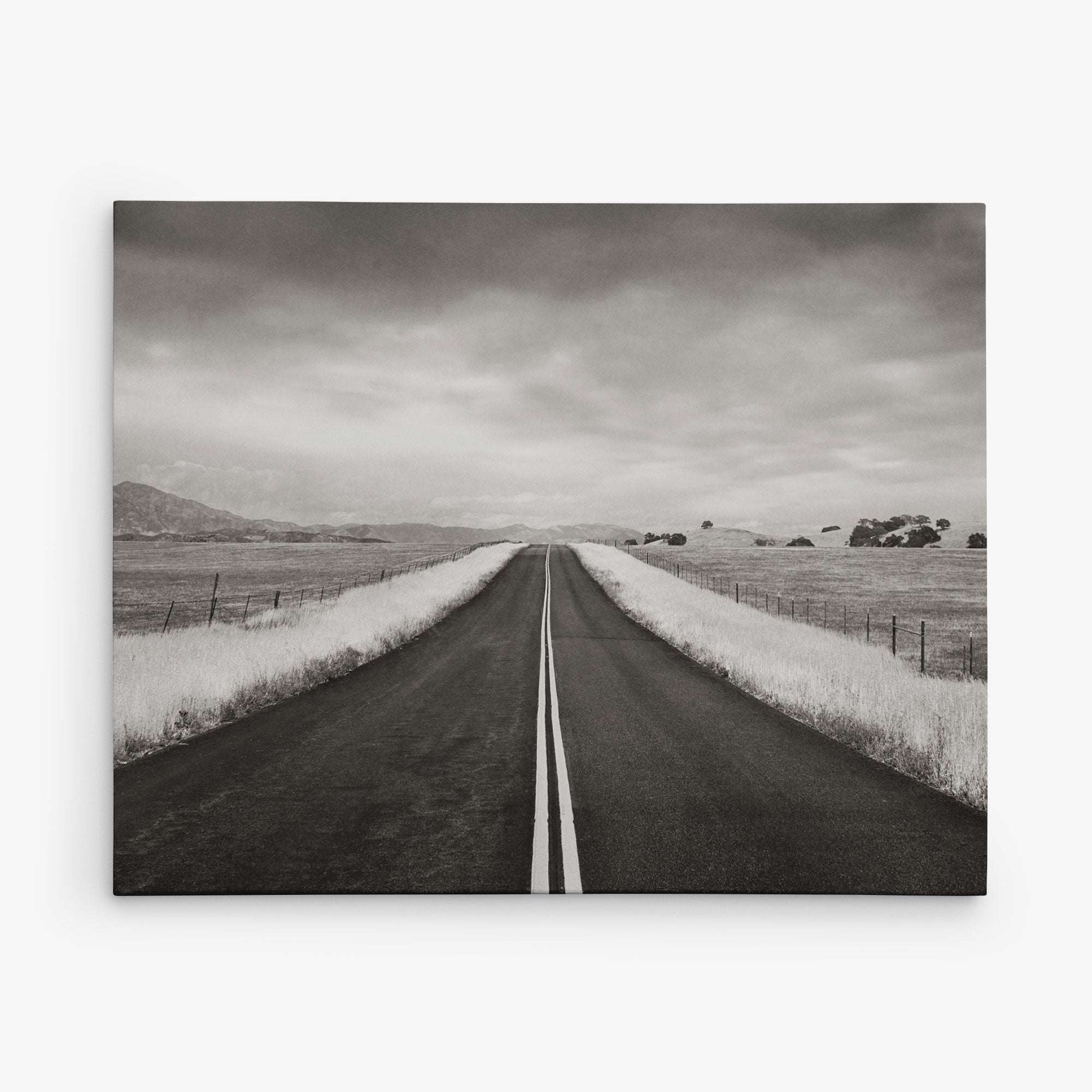 The Offley Green 8x10 Rustic Canvas Print features a black and white photo of an empty road stretching into the distance, bordered by grassy fields under a cloudy sky, creating depth and solitude in the landscape. Choose from 10+ designs.