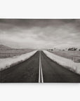 The Offley Green 8x10 Rustic Canvas Print features a black and white photo of an empty road stretching into the distance, bordered by grassy fields under a cloudy sky, creating depth and solitude in the landscape. Choose from 10+ designs.