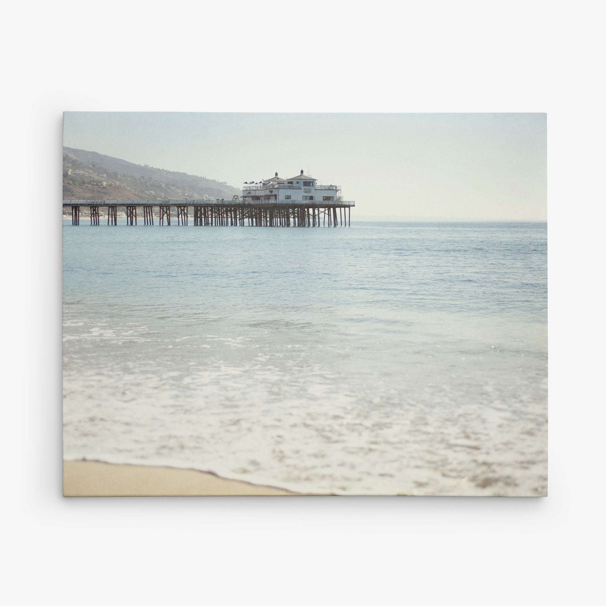 A serene seascape featuring a calm ocean gently lapping against a sandy shore, with Offley Green's Malibu Pier Wall Art extending into the water under a hazy sky.