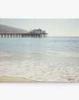 A serene seascape featuring a calm ocean gently lapping against a sandy shore, with Offley Green's Malibu Pier Wall Art extending into the water under a hazy sky.