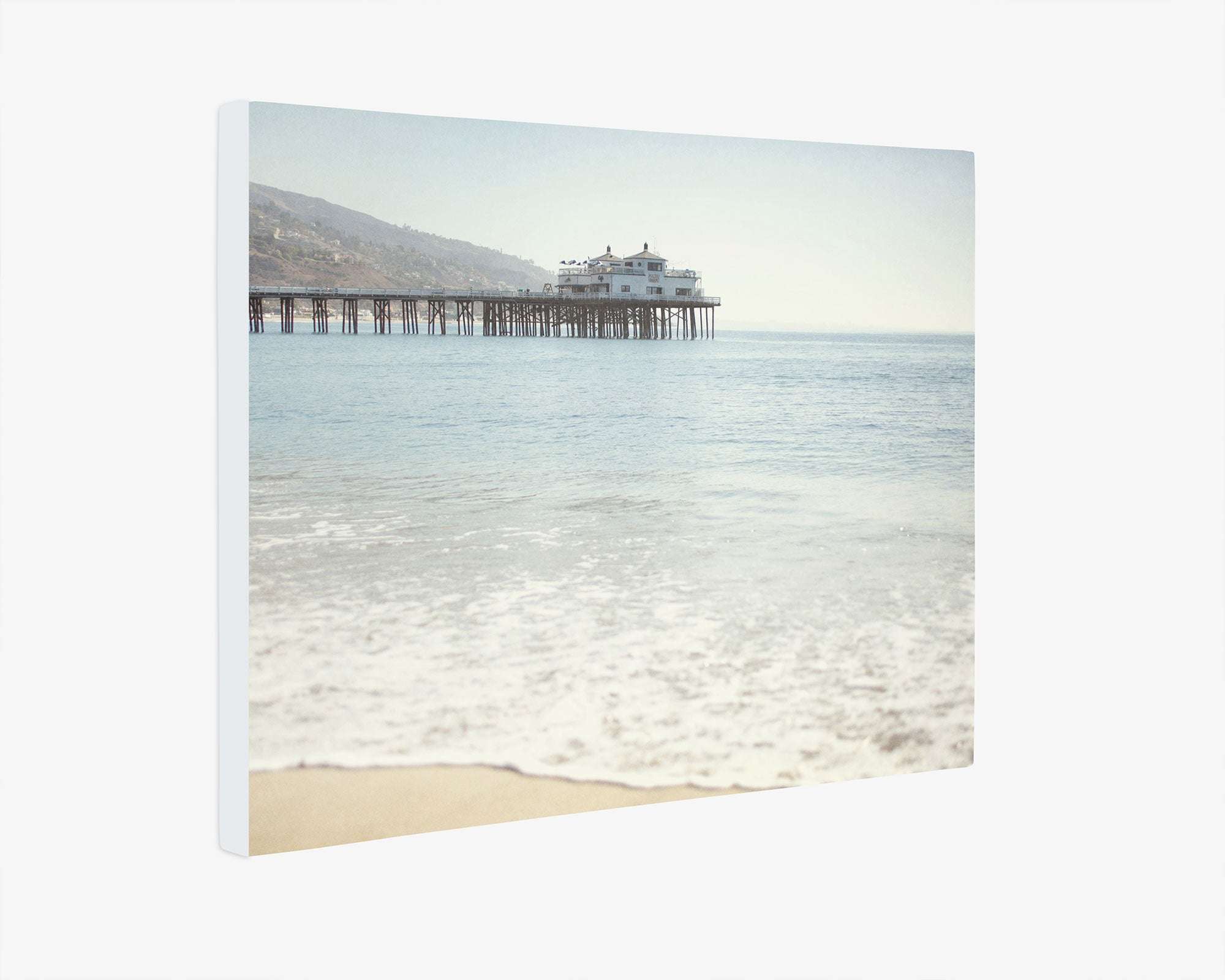 Canvas print of Malibu Pier Wall Art, 'Malibu Pier' by Offley Green extending into the ocean, under a hazy sky, with a building at the end and gentle waves lapping at a sandy beach.