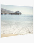Canvas print of Malibu Pier Wall Art, 'Malibu Pier' by Offley Green extending into the ocean, under a hazy sky, with a building at the end and gentle waves lapping at a sandy beach.