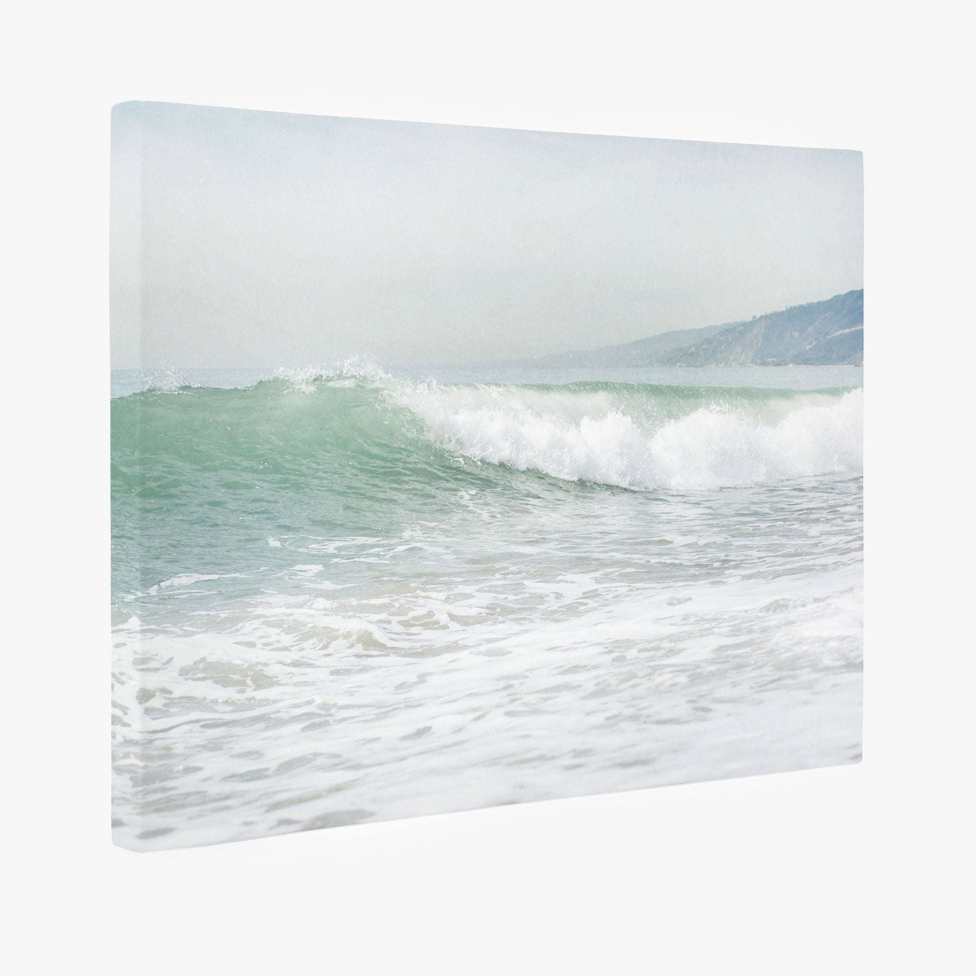 The Coastal Ocean Canvas Waves Wall Art, 'Breaking Surf' by Offley Green depicts a serene ocean wave gently crashing near the shore under coastal sunshine. Distant hills on the right add depth, creating a calm and peaceful scene perfect for relaxation. This beach-themed wall art is crafted on premium artist-grade canvas for lasting quality.