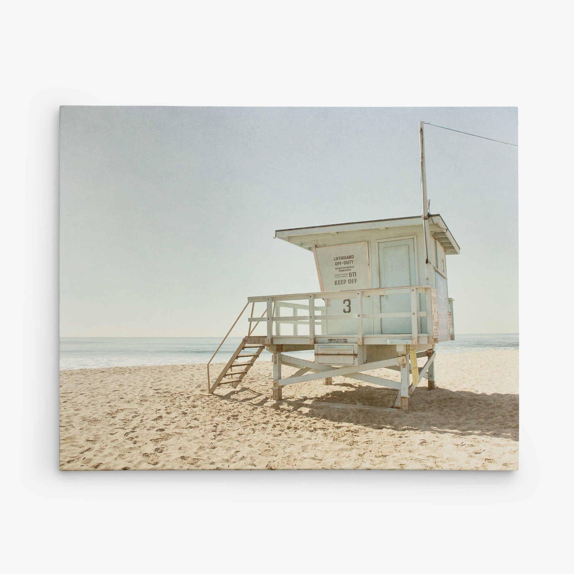 A solitary lifeguard tower stands on a sandy beach under a clear sky. The structure, painted in a weathered white, overlooks the calm ocean. The steps leading up to the platform are empty, and there are no people around, creating a tranquil scene reminiscent of Offley Green's 16x20 California Canvas Print (Choose from 10+ Designs).
