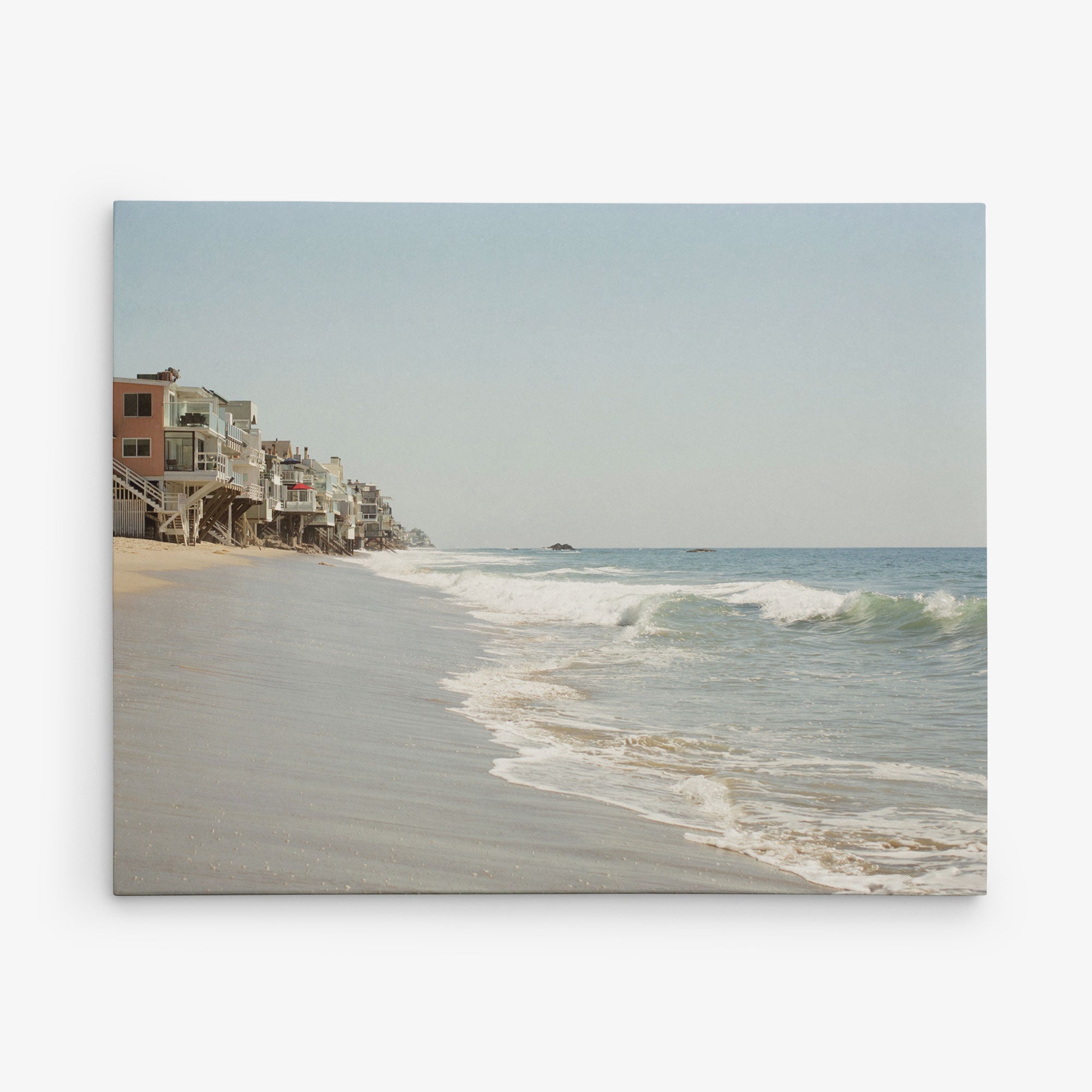 A serene beach scene with gentle waves washing ashore. To the left, colorful beachfront houses on stilts line the sandy coast. The clear blue sky contributes to the peaceful atmosphere, making it an ideal subject for Offley Green's 16x20 California Canvas Print (Choose from 10+ Designs).