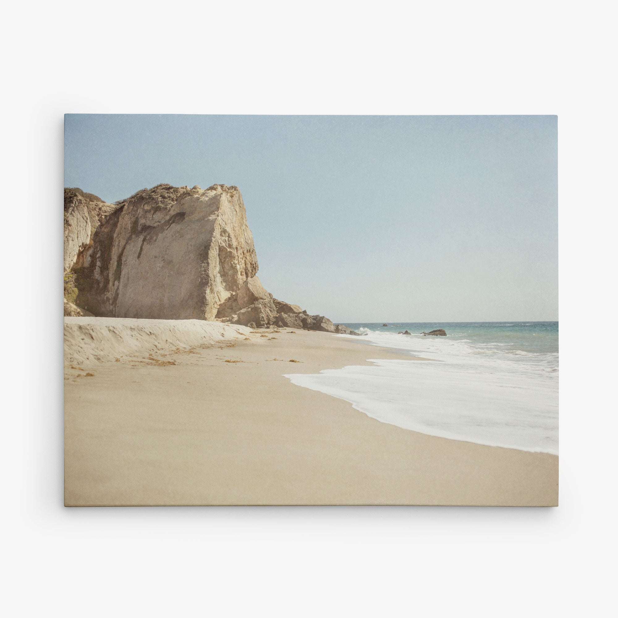 A serene beach scene at Point Dume with a large rocky cliff on the left side. The sandy shore is expansive and meets gentle ocean waves under a clear blue sky. The sea stretches to the horizon, creating a tranquil and picturesque seaside atmosphere, perfect for California Malibu Canvas Wall Art, 'Point Dume' by Offley Green.