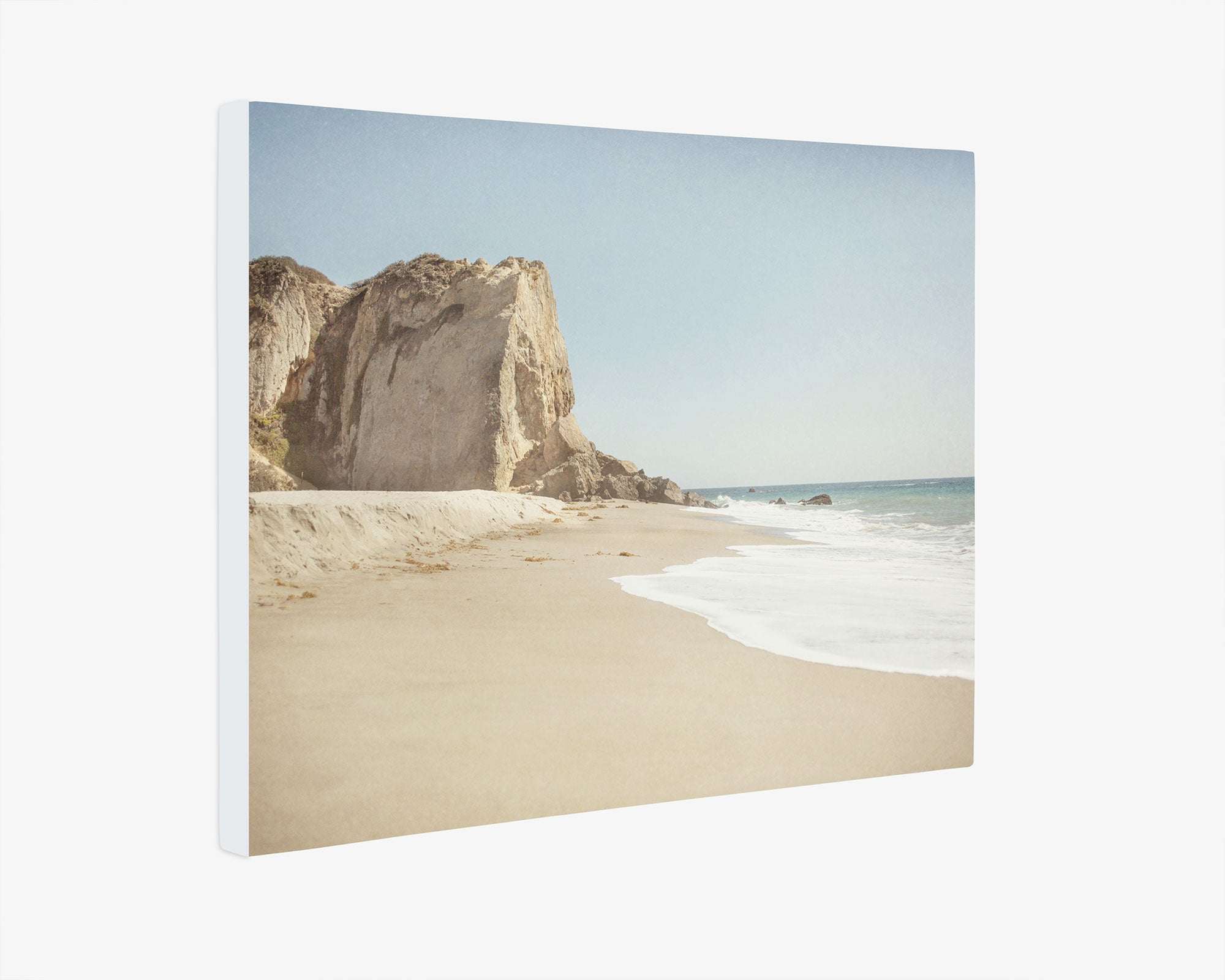 A serene beach scene with waves gently lapping at the shore and a large rocky cliff in the background under a clear blue sky, beautifully capturing Point Dume. This California Malibu Canvas Wall Art, 'Point Dume' by Offley Green is perfect as a canvas gallery wrap to bring tranquility into any room.