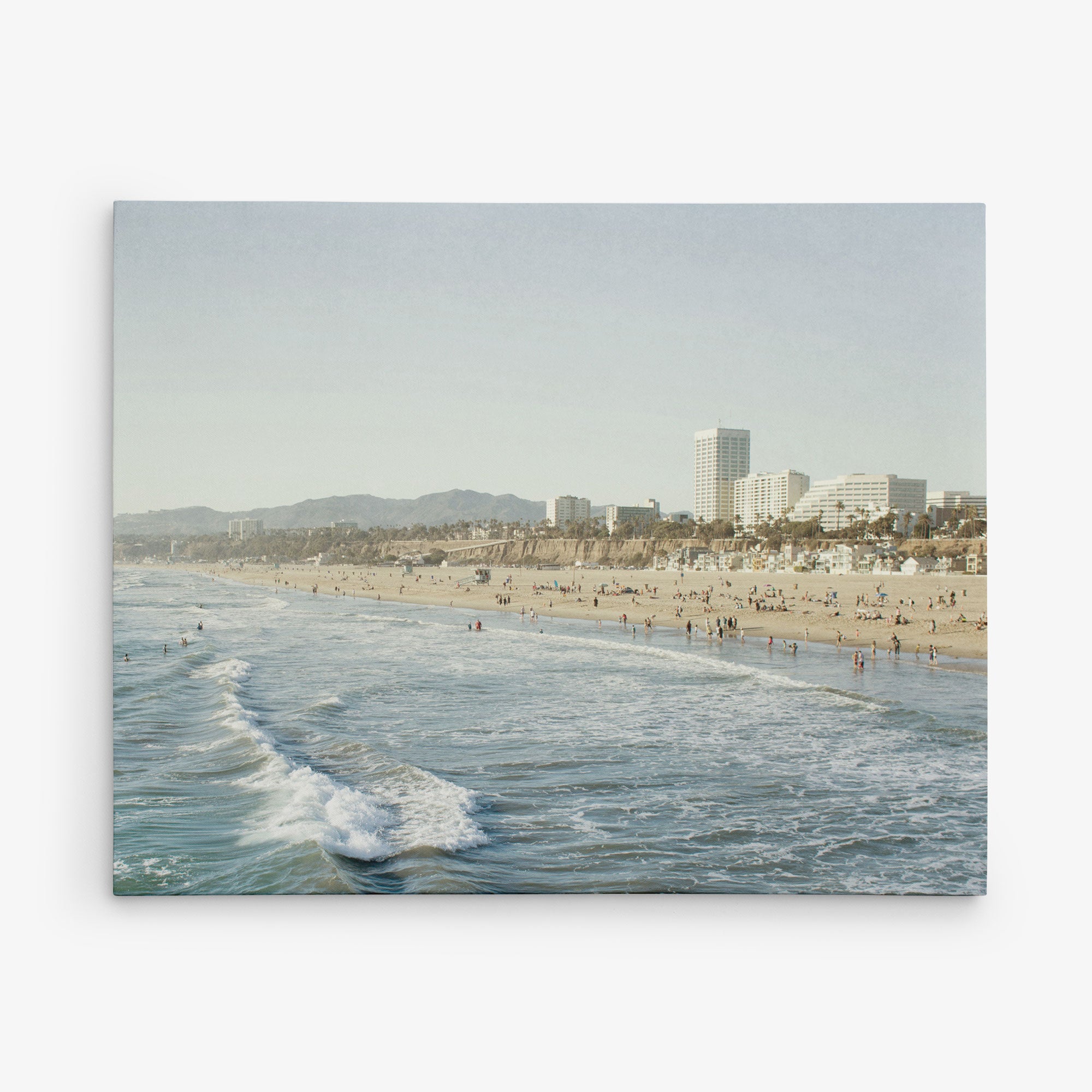 The Santa Monica Canvas Wall Art, 'Santa Monica Seaside' by Offley Green captures a bustling coastal scene with light sand and gentle waves. Numerous people are visible enjoying the shore. In the background, tall buildings rise against hills and a clear, slightly hazy sky, reminiscent of a day at Santa Monica Pier. Perfect for canvas gallery wrap.
