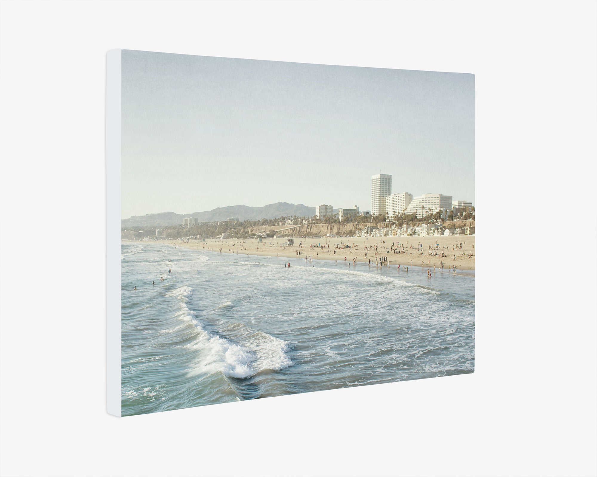 The Offley Green Santa Monica Canvas Wall Art, 'Santa Monica Seaside' showcases a serene beach scene with gentle waves rolling onto the sandy shore. The background features buildings, possibly part of a coastal city like Santa Monica Pier, and is lined with distant mountains under a clear sky. Perfect as beach-themed wall art.